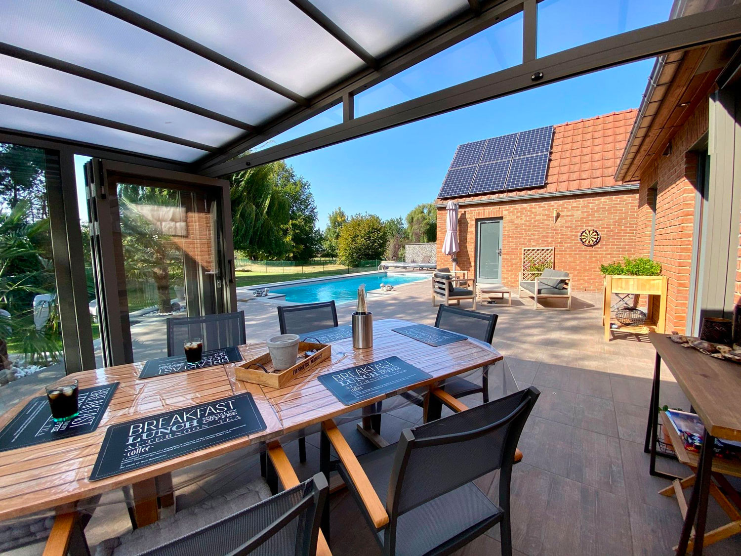 Abri de terrasse rétractable ouvert sur le reste de la terrasse et piscine. Sous l'abri il y a une table en bois dressée pour le petit déjeuner et des chaises