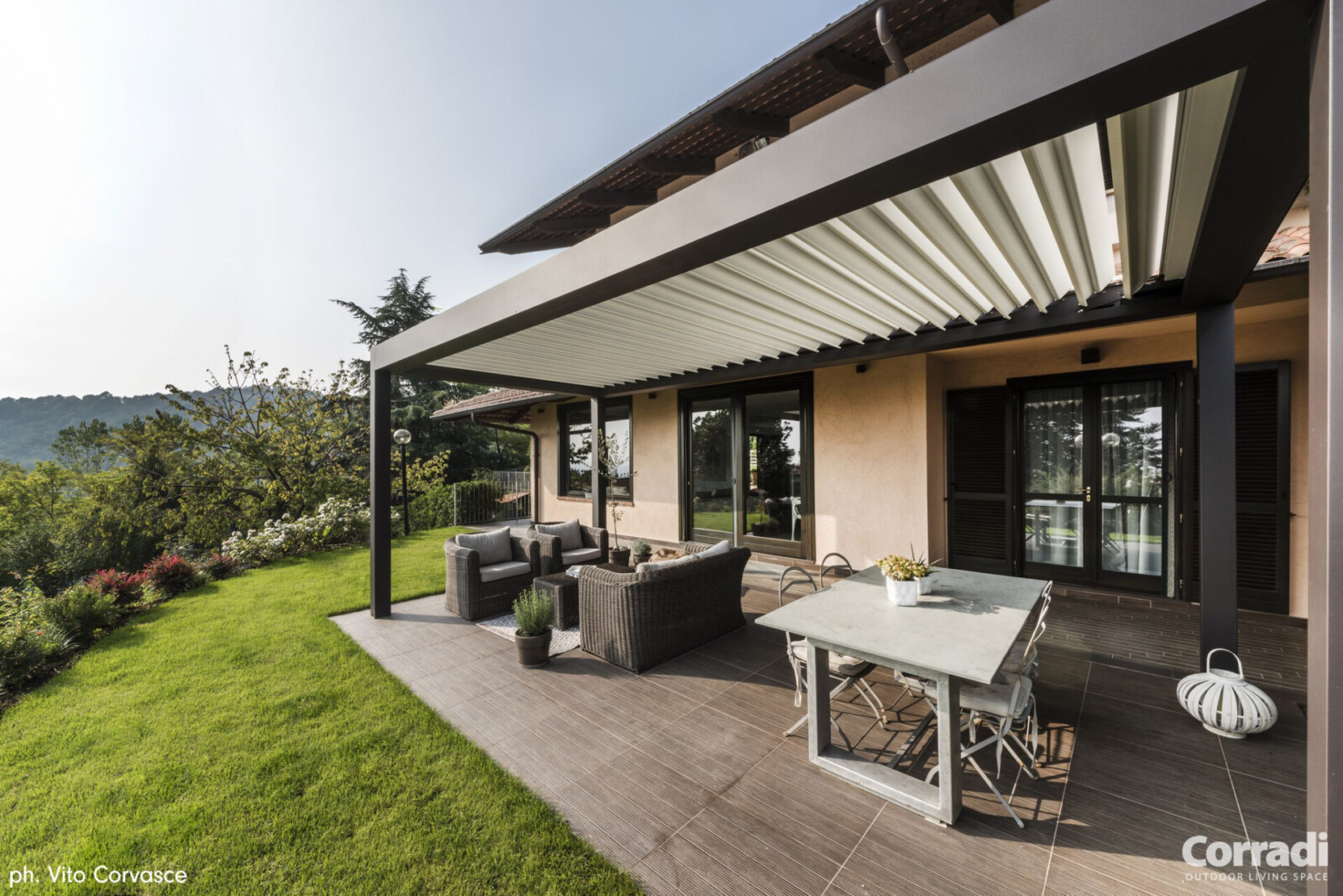 Pergola bioclimatique adossée au mur d'une maison, abrite un salon de jardin extérieur et donne un accès direct au jardin