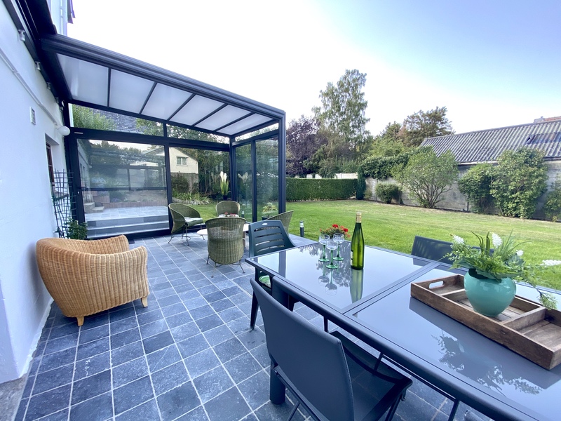 Véranda rétractable sur une terrasse carrelée à ras du jardin, composée d'une table en verre et des chaises en osier