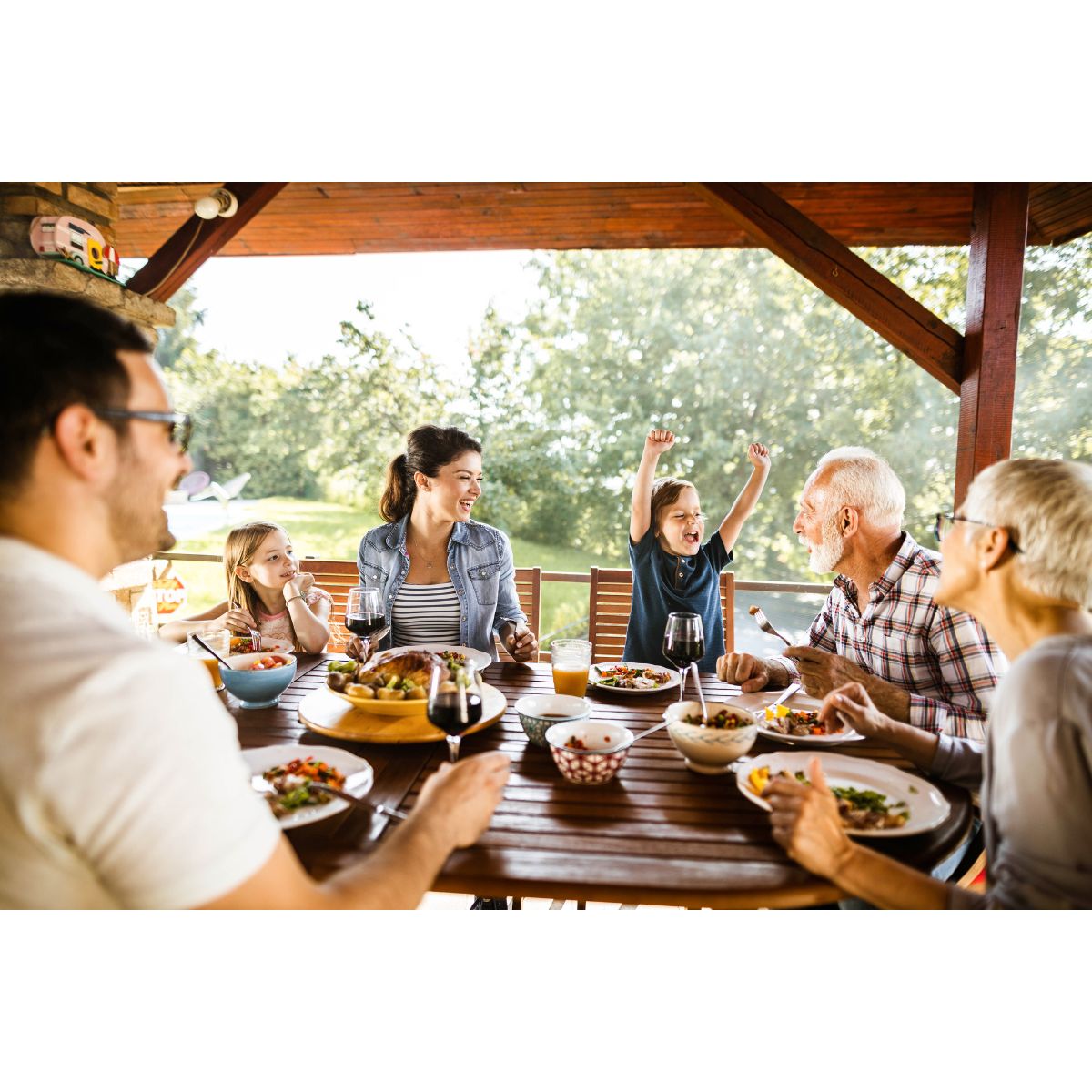 Une famille composée d'enfants, de leurs parents et de leurs grands-parents prend un repas sur la terrasse extérieur protégée par une protection de terrasse