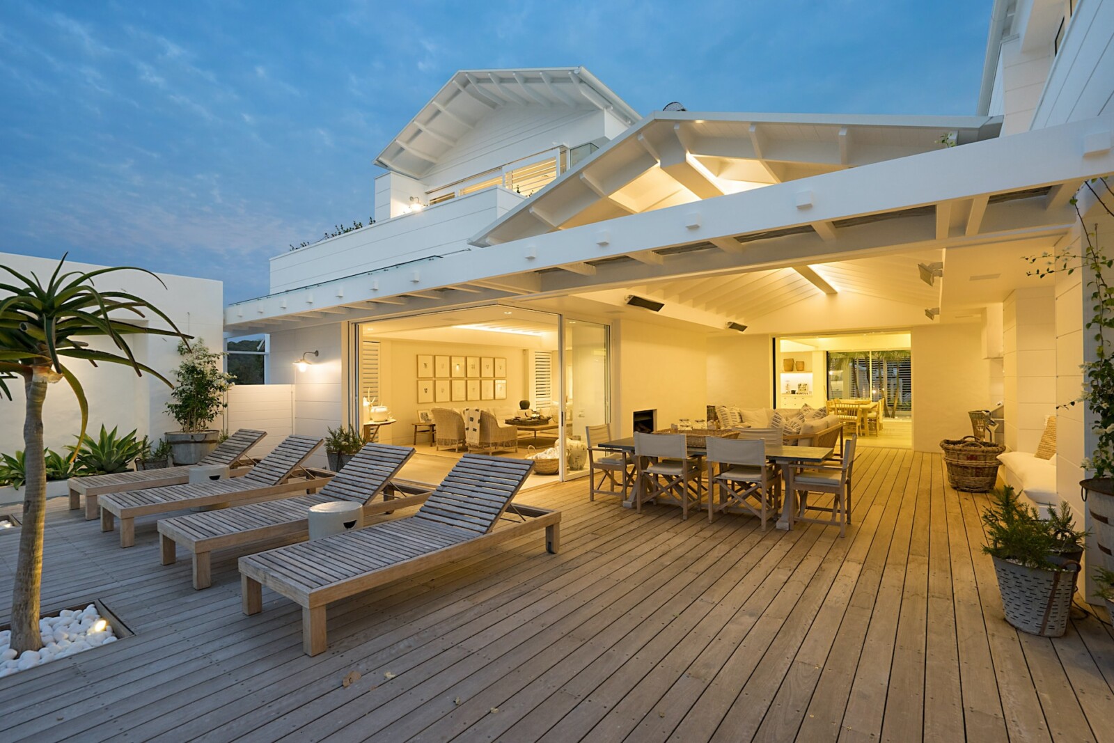 Patio blanc pour aménager la terrasse extérieur en bois, composée de transats en bois, d'une table à manger et d'un palmier.