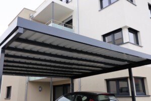 Carport en aluminium gris avec toit en polycarbonate ondulé, installé devant un bâtiment résidentiel beige avec des fenêtres à cadre noir et un balcon en verre.