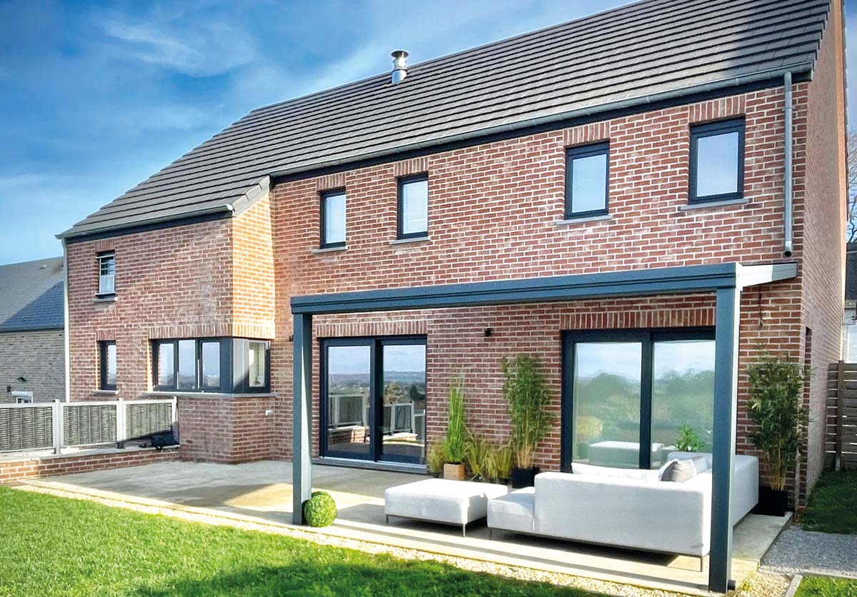 Pergola toit fixe ouverte accolée à une maison aux briques rouges. Il y a un fauteuil de terrasse gris et des plantes vertes