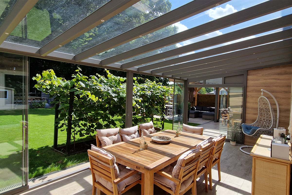 Pergola vitrée fermée avec tables et chaises en bois et une vue sur le jardin et une vigne