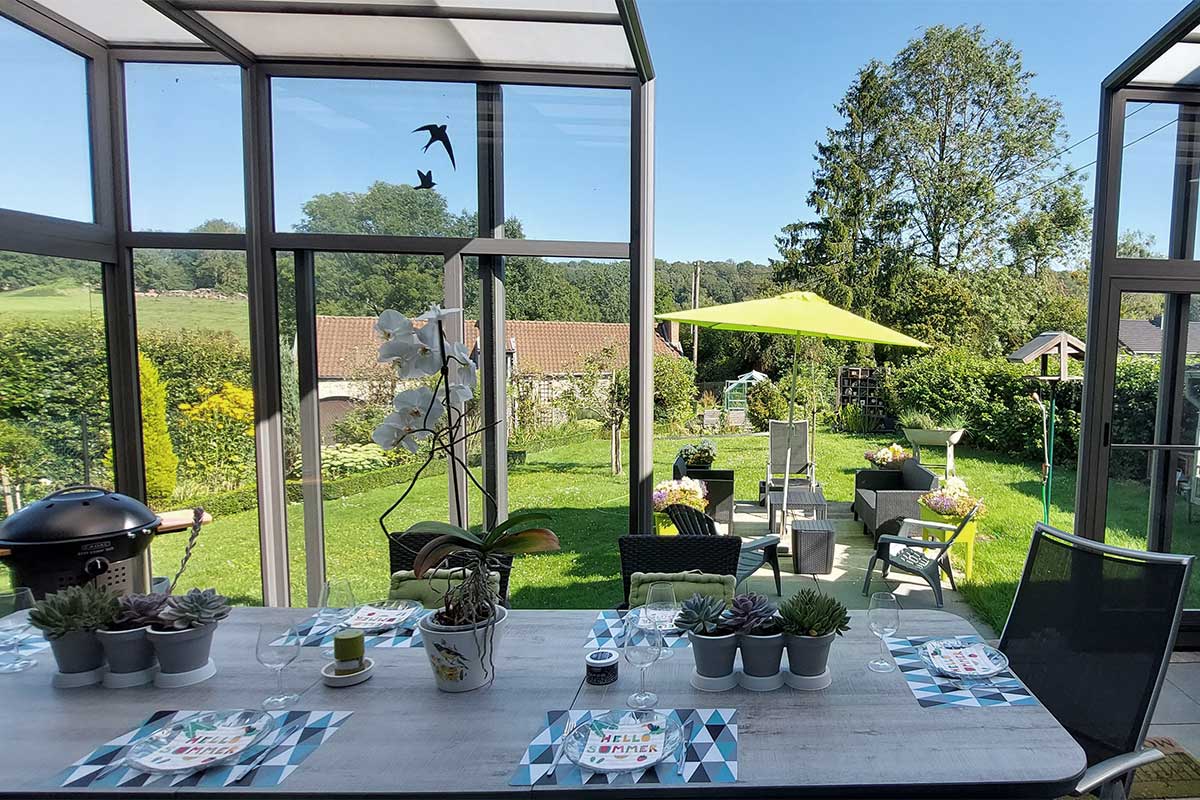 Abri de terrasse rétractable avec une vue sur le jardin, un parasol vert, un barbecue, une table dressée sur le thème de l'été