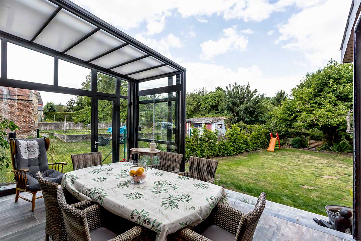 Abri de terrasse rétractable. Il y a une table avec une nappe, des chaises et une vue sur le jardin avec un toboggan pour enfant