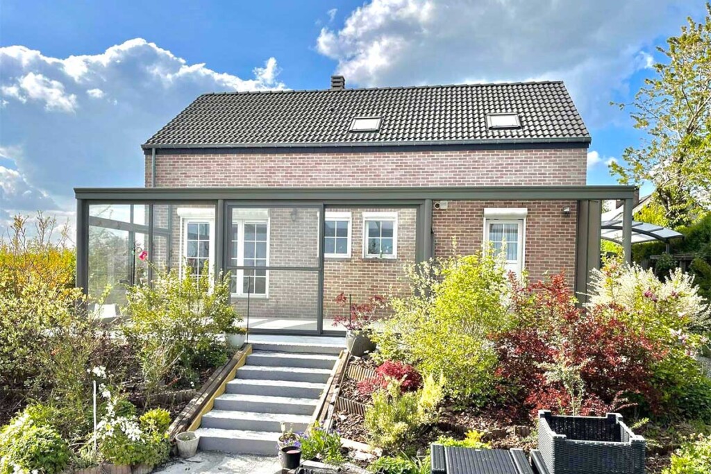 Pergola vitrée accolée à une maison à briques beiges et rouges et sur une terrasse qui donne accès au jardin avec un petit escalier