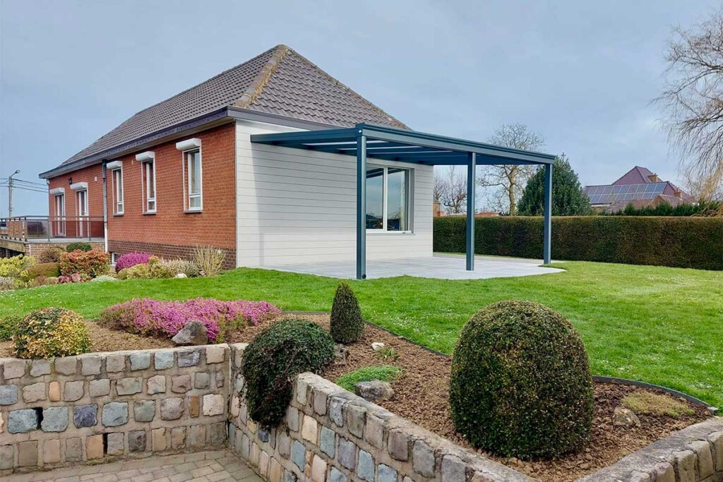 Pergola ouverte accolée à un mur de maison en bois blanc et une vue sur un jardin fleuri