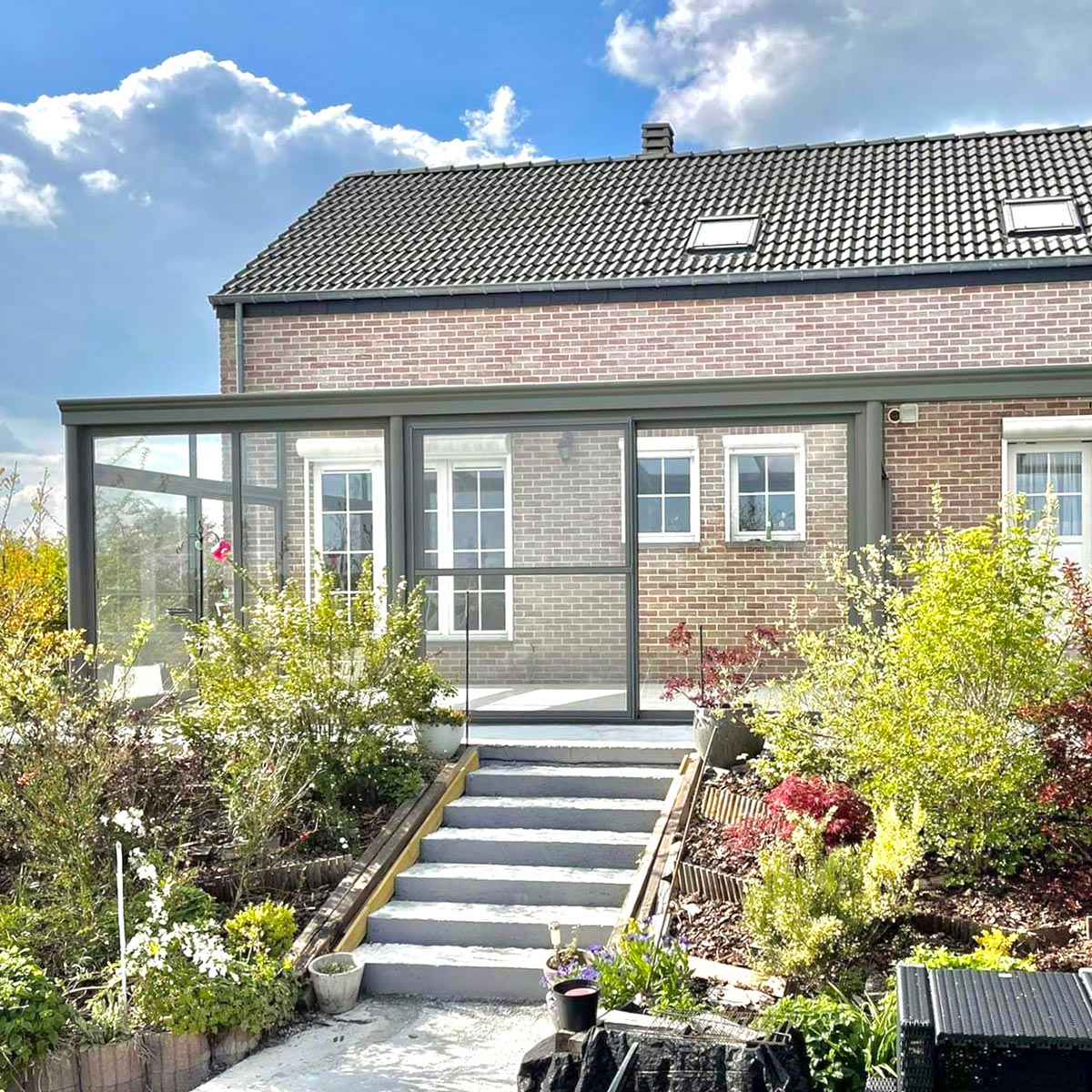 Pergola vitrée accolée à une maison à briques beiges et rouges et sur une terrasse qui donne accès au jardin avec un petit escalier