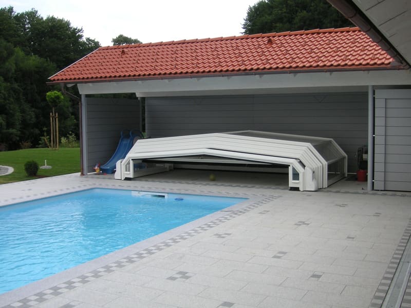 Abri de piscine bas amovible blanc pour une piscine entourée de carrelages gris avec un abri en bois gris