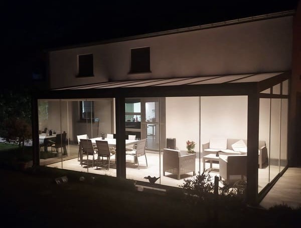 Pergola vitrée accolée à une maison blanche avec une table, des chaises et des fauteuils sous la pergola