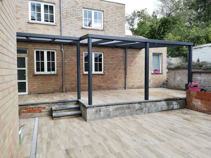 Pergola à toiture fixe adossé à une maison en briques claires. La couleur de la pergola est gris anthracite.