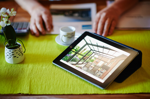 visualisation projet verandair Une tablette est posée sur un chemin de table vert. Elle affiche la photo d'un intérieur lumineux avec des baies vitrées. À côté, une tasse de café sur une soucoupe et un pot avec une plante verte