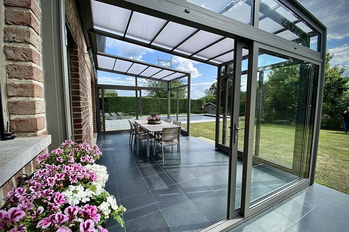 Installation d'une pergola Verandair équipée de parois coulissantes en verre finition gris anthracite. Les fenêtres sont ornées de fleurs blanches et roses.