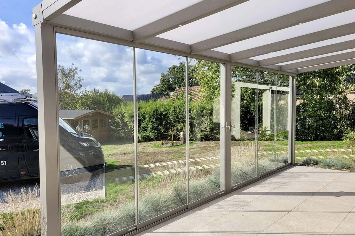 Pergola moderne de chez Verandair avec un toit en lames blanches et des montants verticaux en aluminium. Elle est composée de parois de verre transparentes coulissantes qui donne une ouverture sur le jardin bien entretenu