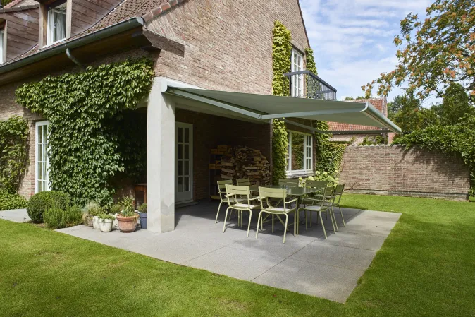 Véranda rétractable sur une terrasse carrelée à ras du jardin, composée d'une table en verre et des chaises en osier